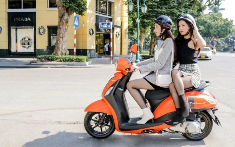 Hai người ngồi trên xe tay ga màu cam ở góc phố, đội mũ bảo hiểm và mặc trang phục thời trang.