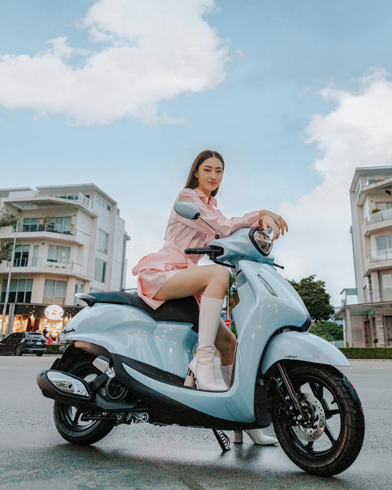 Người ngồi trên xe tay ga màu xanh nhạt, mặc trang phục màu hồng, nền là tòa nhà và bầu trời xanh.