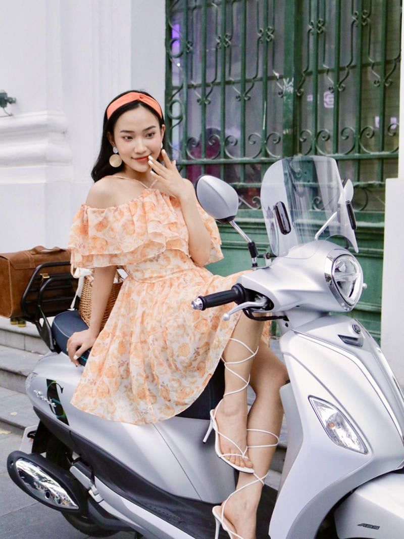 Người phụ nữ ngồi trên xe tay ga màu bạc, mặc váy hoa cam, đeo băng đô đỏ, phía sau là cửa sổ sắt.