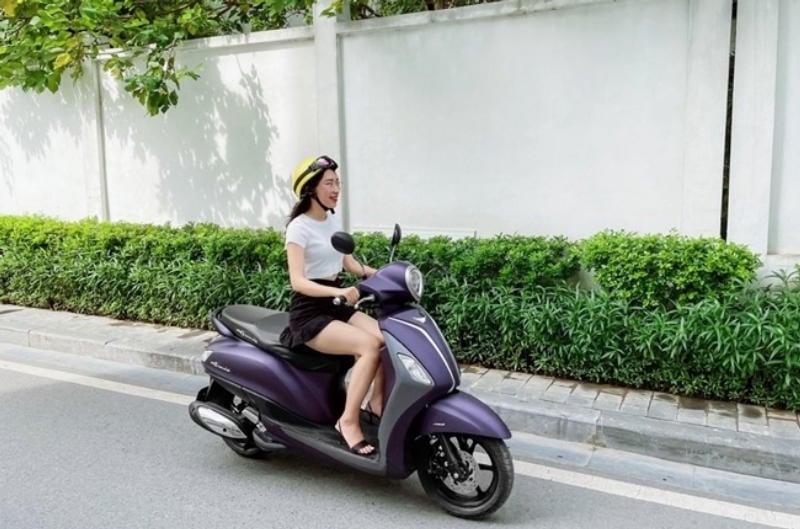 Người phụ nữ đội mũ bảo hiểm màu vàng, lái xe máy màu tím trên con đường cạnh hàng rào trắng và hàng cây xanh.