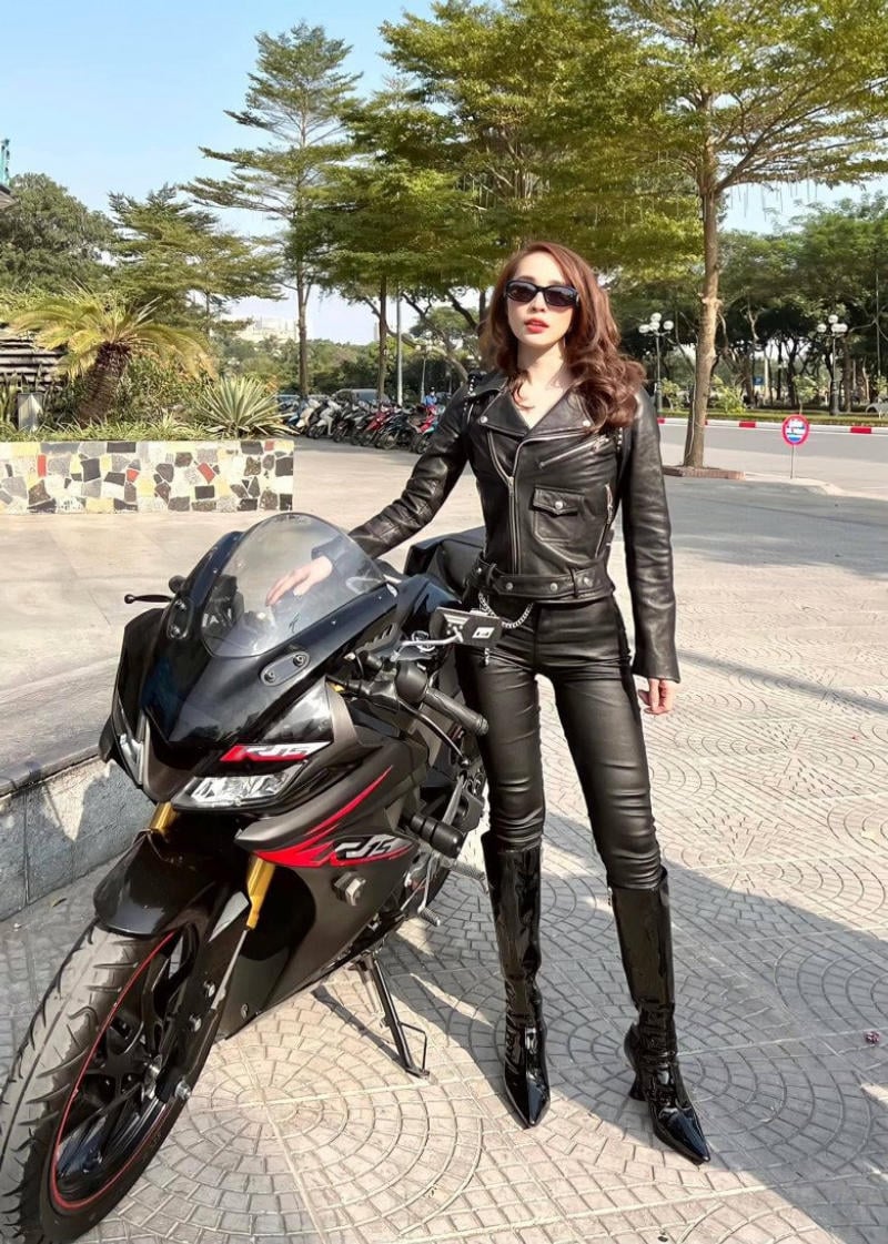 Người mặc đồ da đen đứng cạnh xe máy thể thao trên vỉa hè có cây xanh và xe máy khác phía sau.