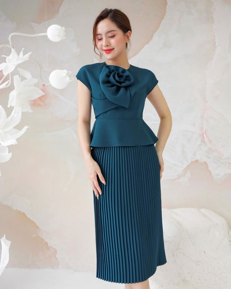 Người phụ nữ mặc váy xanh đậm với chi tiết hoa lớn, đứng trước nền tường màu nhạt.