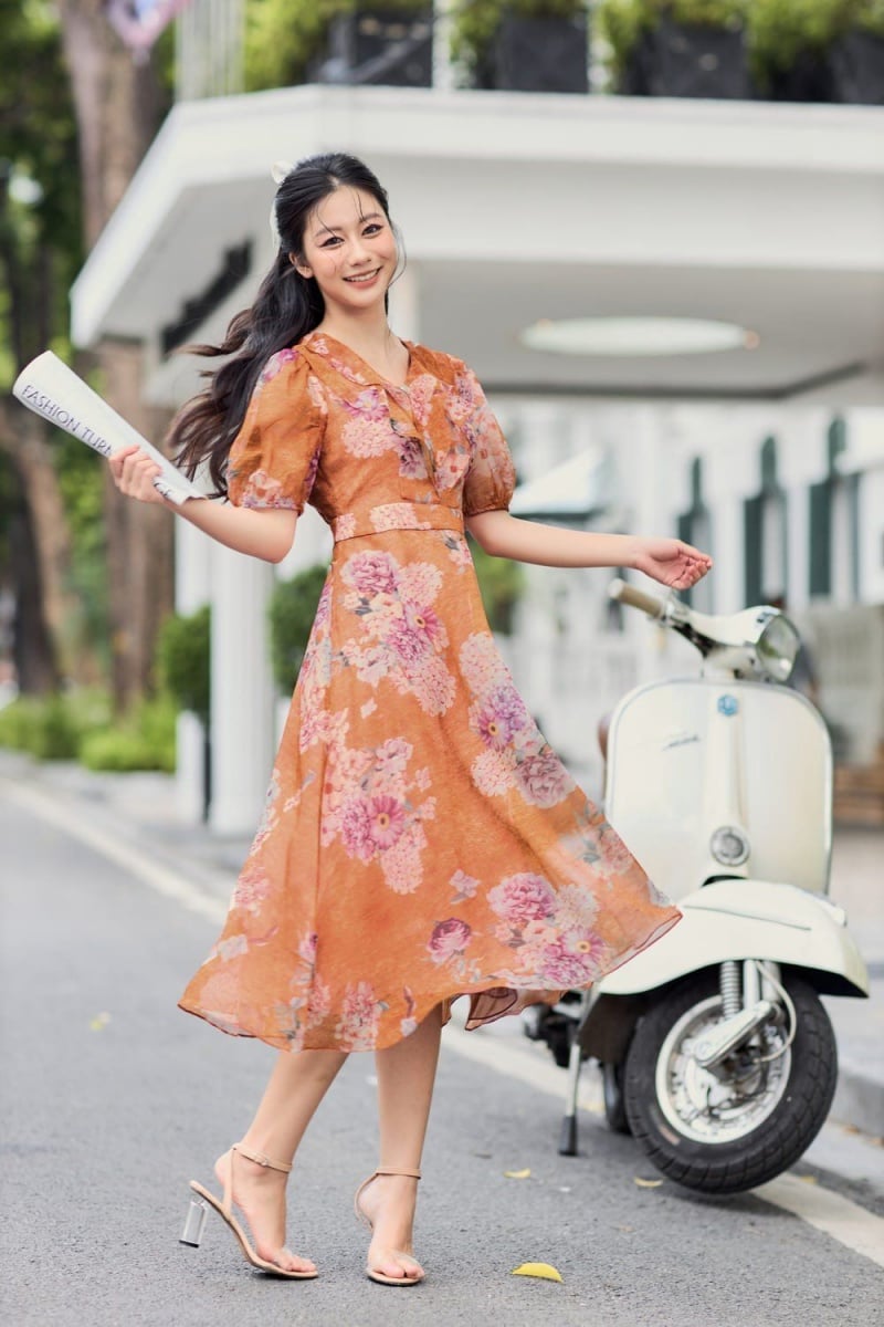 Người phụ nữ mặc váy hoa cam đứng gần xe máy cổ điển, cầm tờ báo trên đường phố.