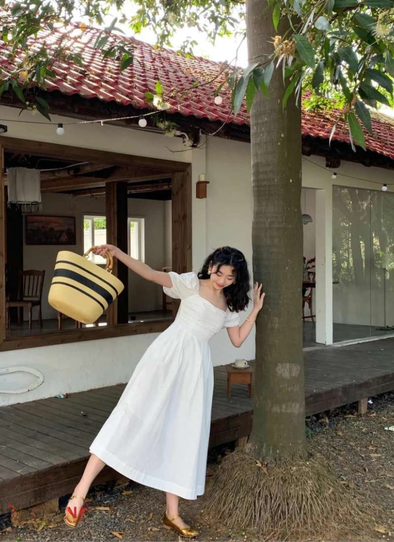 Người mặc váy trắng đứng cạnh cây, cầm túi xách sọc, trước một ngôi nhà mái ngói đỏ.