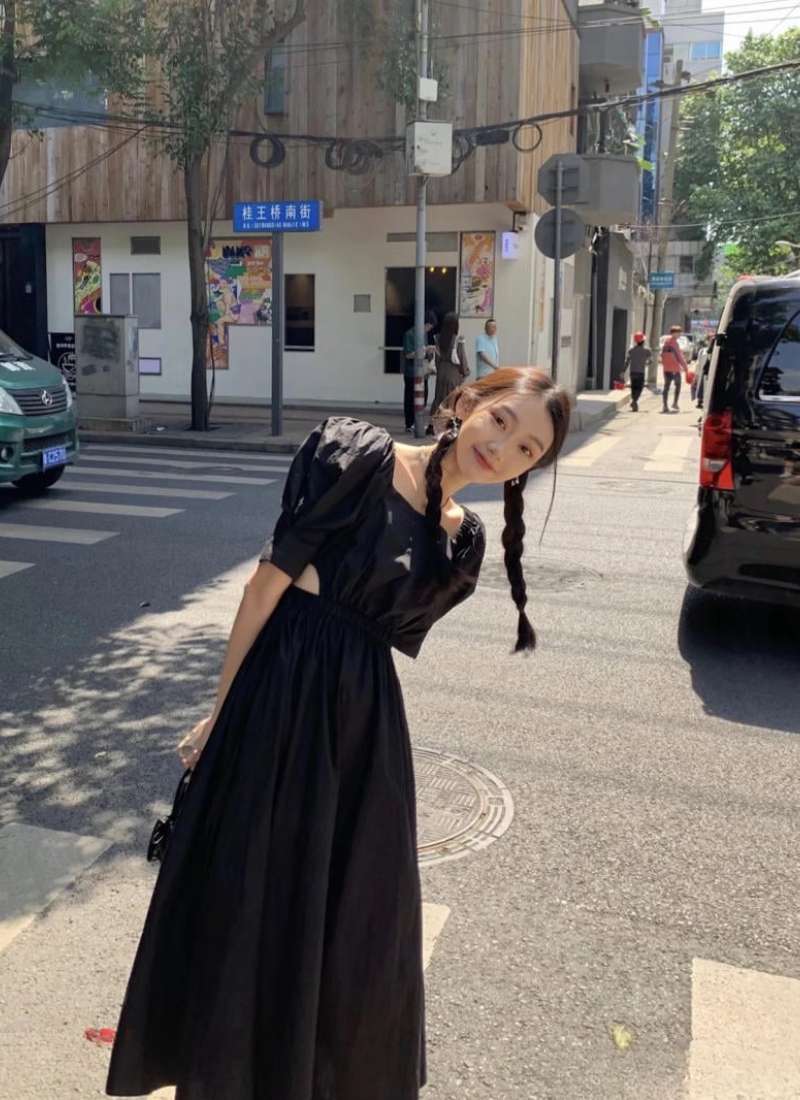Người phụ nữ mặc váy đen đứng trên vỉa hè, phía sau là đường phố và tòa nhà.