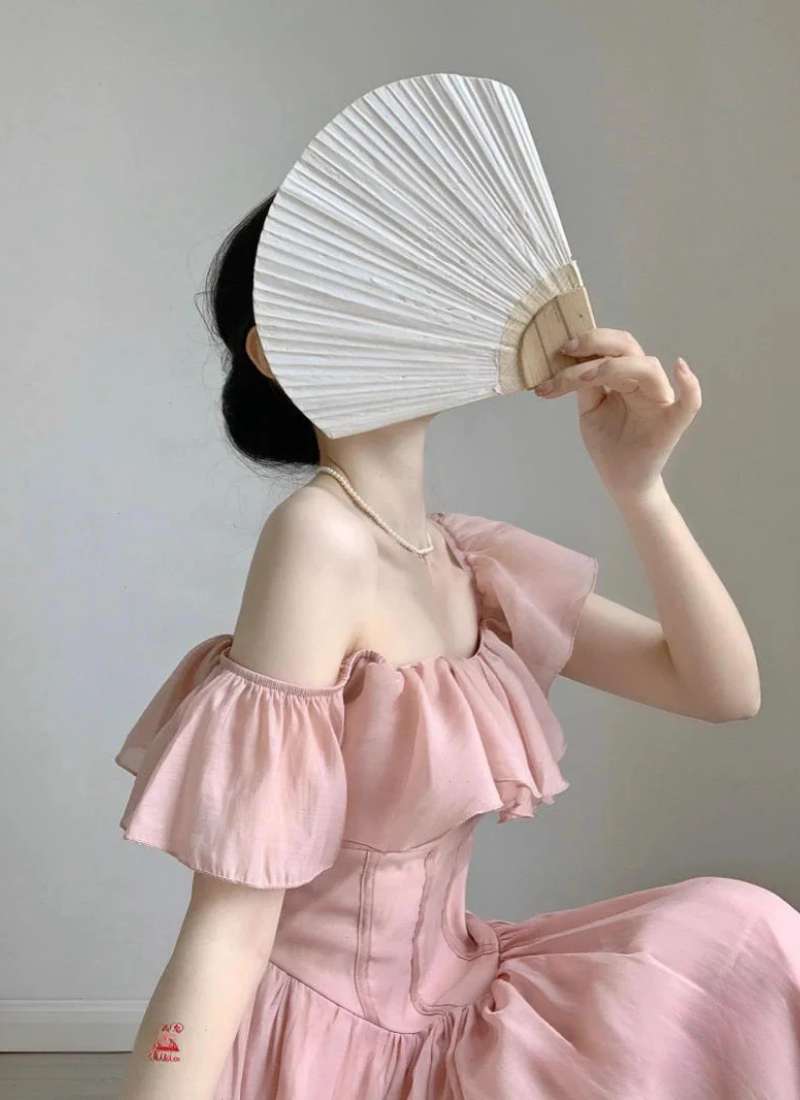 Người mặc váy hồng cầm quạt trắng che mặt, ngồi nghiêng trong không gian tối giản.