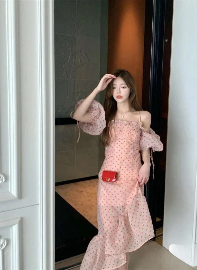 Người mặc váy chấm bi màu hồng đứng trong không gian nội thất, cầm tay lên tóc, nhìn về phía trước.