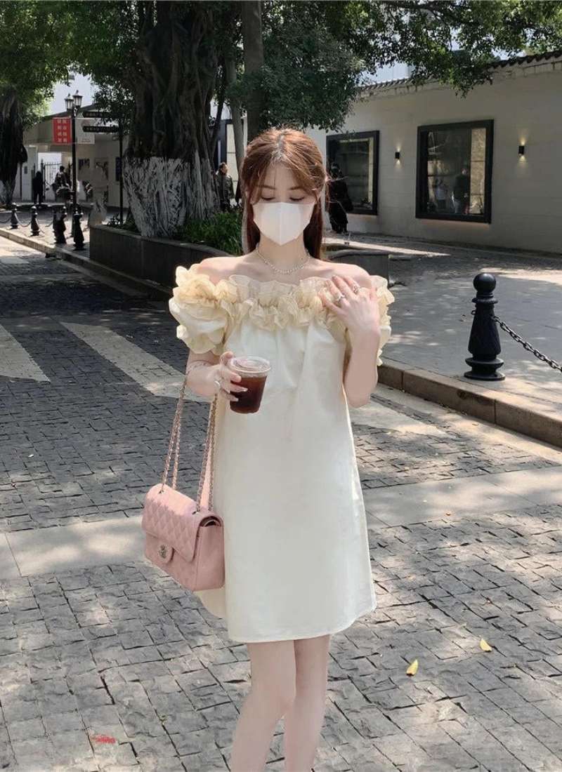 Người phụ nữ mặc váy trắng, đeo khẩu trang, cầm ly nước và túi xách trên phố, bên cạnh là cây xanh và tòa nhà.