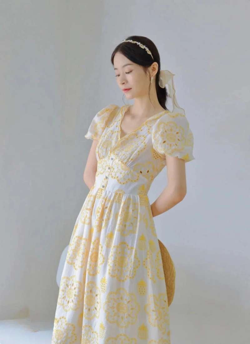 Người phụ nữ mặc váy hoa vàng trắng, đứng nghiêng, nền trắng, tóc cột và đeo băng đô hoa.
