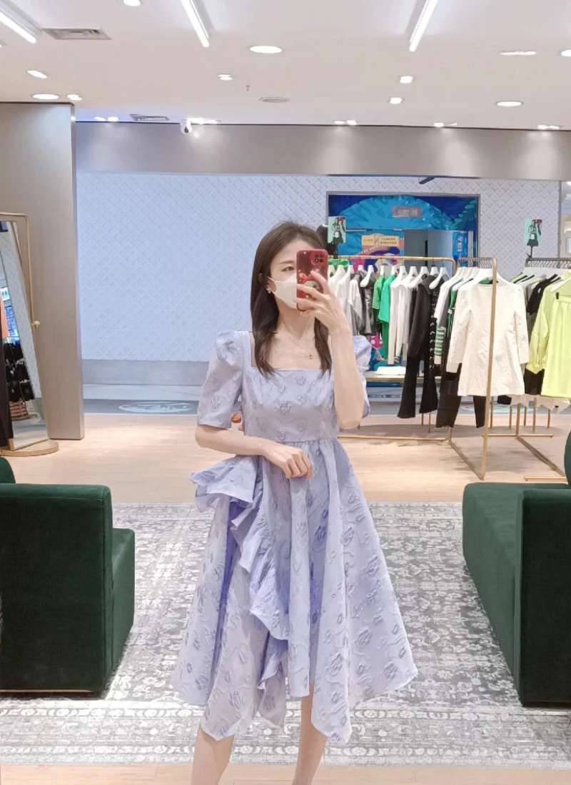 Người phụ nữ chụp ảnh tự sướng trong gương, mặc váy màu tím nhạt, đứng trong một cửa hàng quần áo.