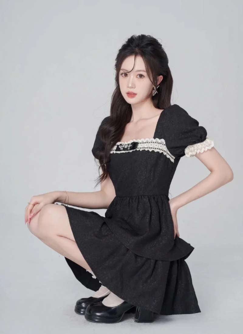 Người mẫu ngồi, mặc váy đen với viền ren trắng, tóc dài, nền màu xám nhạt.