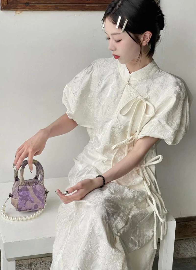 Người mặc váy trắng, cầm túi xách nhỏ màu tím, ngồi trên ghế, tóc buộc gọn với kẹp tóc trắng.