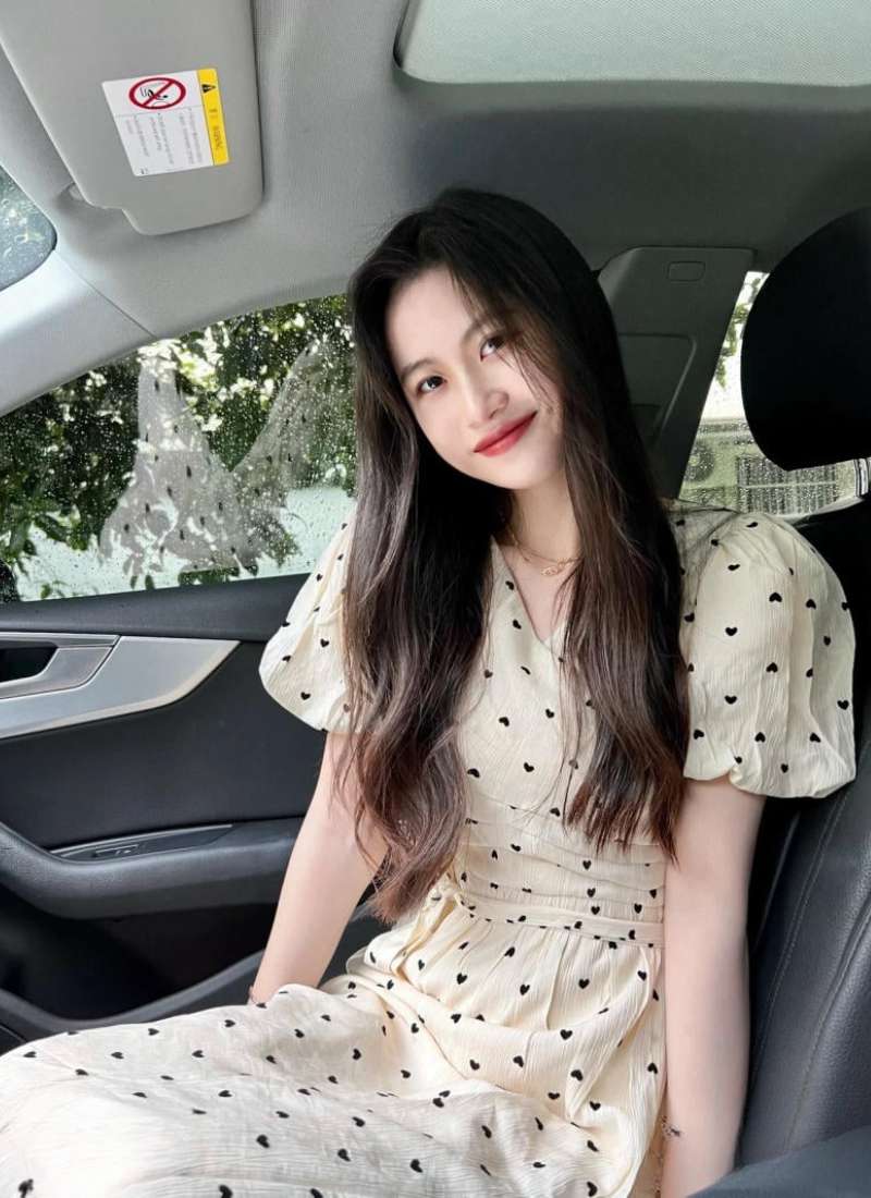Người phụ nữ ngồi trong xe hơi, mặc váy sáng màu với họa tiết trái tim, nhìn về phía máy ảnh.