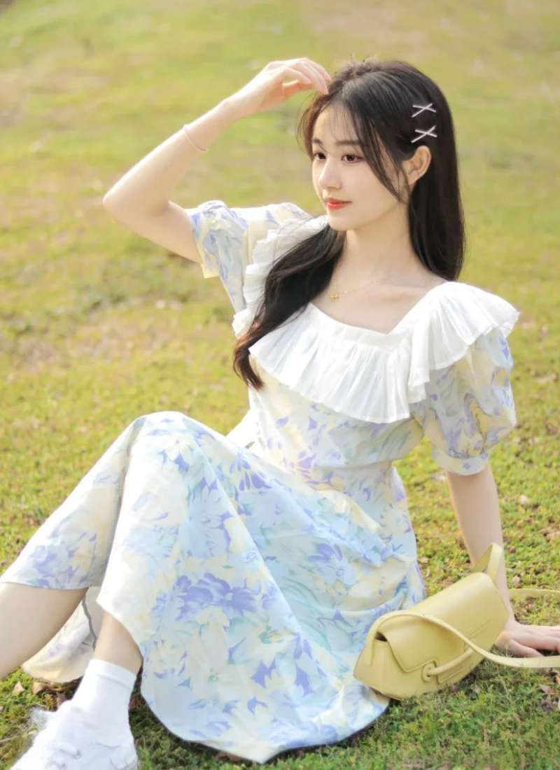 Người phụ nữ mặc váy hoa ngồi trên cỏ, tay chạm tóc, bên cạnh là túi xách màu vàng nhạt.