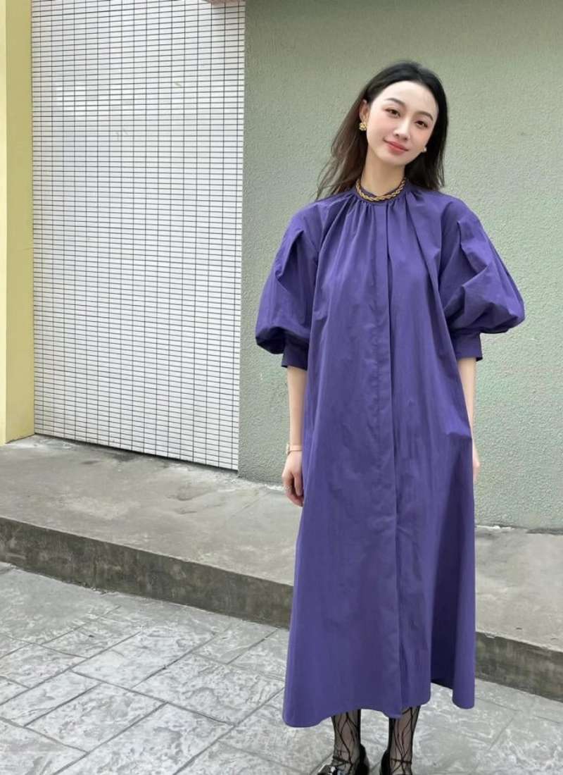 Người phụ nữ mặc váy tím tay phồng đứng ngoài trời, nền là bức tường màu sáng và sàn gạch xám.