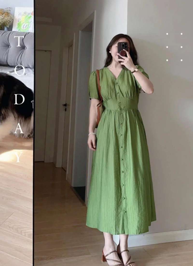Người chụp ảnh qua gương mặc váy xanh lá, cầm điện thoại, đứng trong phòng có sàn gỗ và tường trắng.