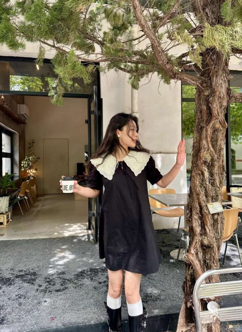 Người phụ nữ mặc váy đen đứng ngoài quán cà phê, tay cầm ly nước, cạnh cây xanh và bàn ghế ngoài trời.