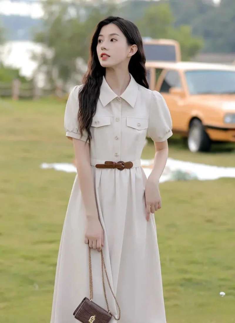 Người phụ nữ mặc váy trắng đứng trên cỏ, phía sau là một chiếc xe hơi màu vàng cam.