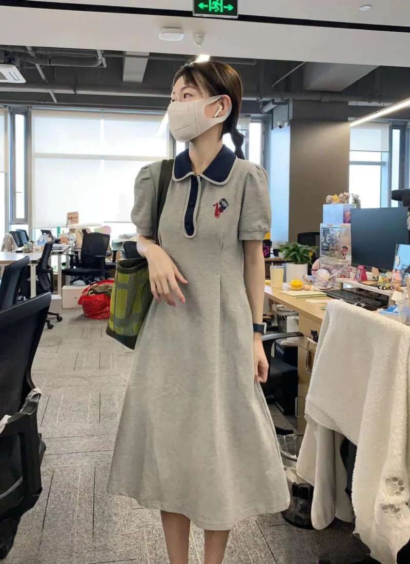 Người đeo khẩu trang đứng trong văn phòng, mặc váy xám và cầm túi, không gian xung quanh có bàn và ghế làm việc.