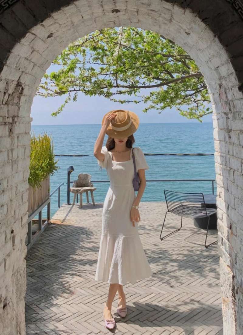 Người mặc váy trắng đội nón đứng dưới cổng vòm, phía sau là biển xanh và cây xanh.