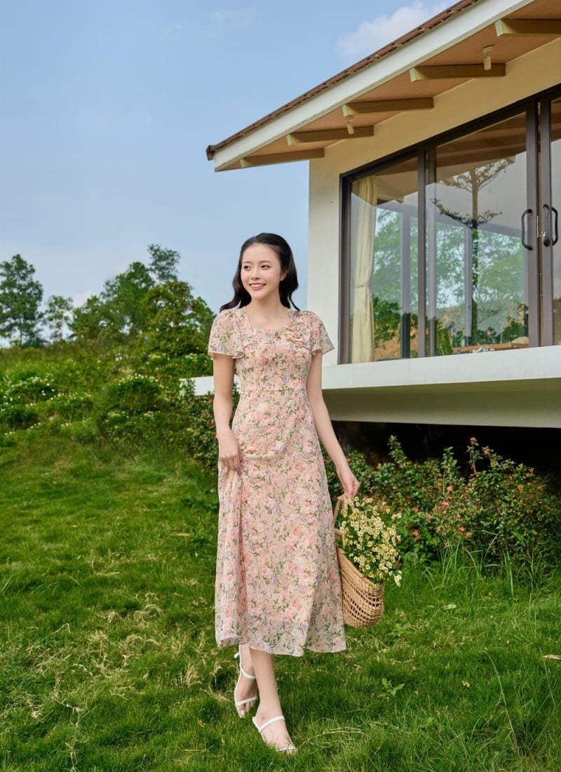 Người phụ nữ mặc váy hoa, đứng ngoài trời gần căn nhà, cầm giỏ hoa, nền cỏ xanh và cây xanh.