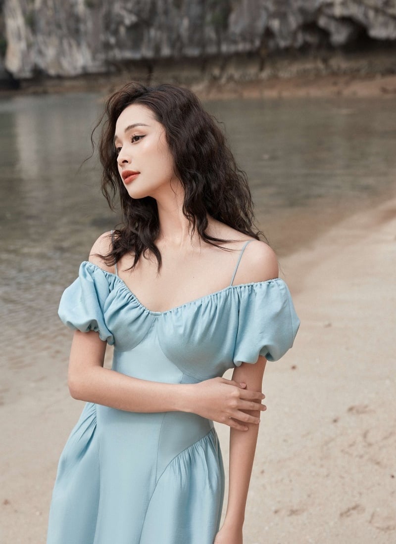 Người phụ nữ mặc váy xanh đứng trên bãi biển với mái tóc xoăn, phông nền là vách đá.