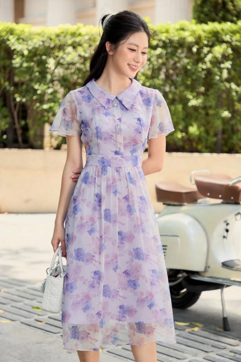 Người phụ nữ mặc váy hoa tím nhạt, tay cầm túi xách trắng, đứng cạnh xe máy cổ điển ngoài trời.