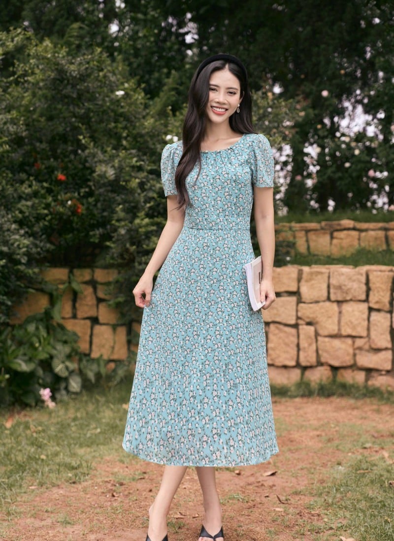 Người phụ nữ mặc váy hoa xanh đứng cầm sách, mỉm cười trước bức tường đá và cây cỏ xanh tươi.