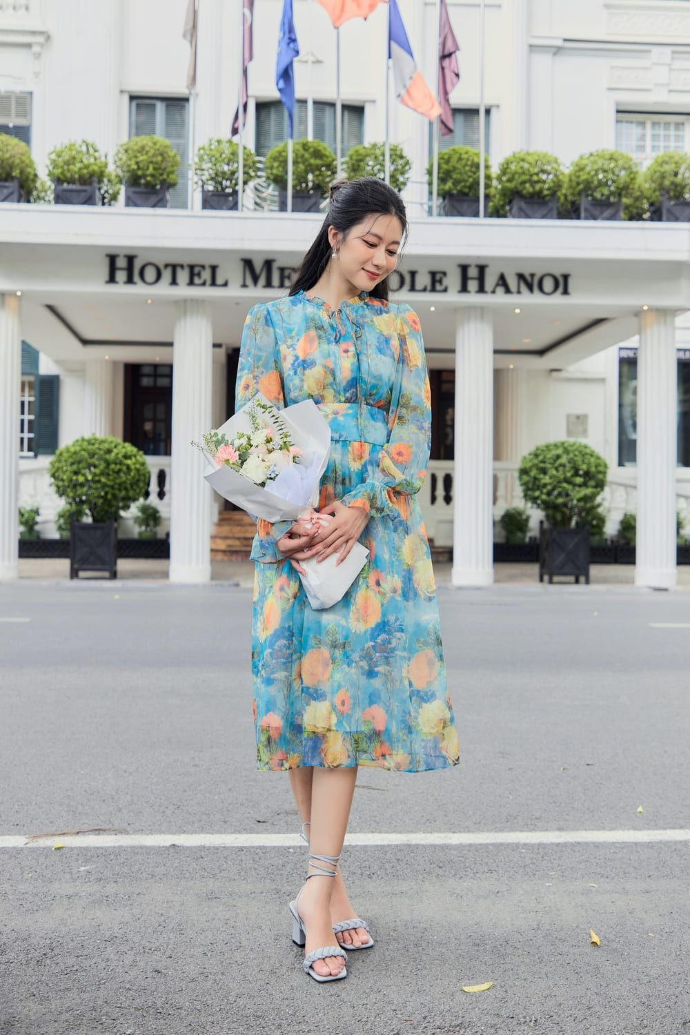 Người phụ nữ mặc váy hoa, cầm bó hoa, đứng trước tòa nhà với biển hiệu "Hotel Metropole Hanoi".