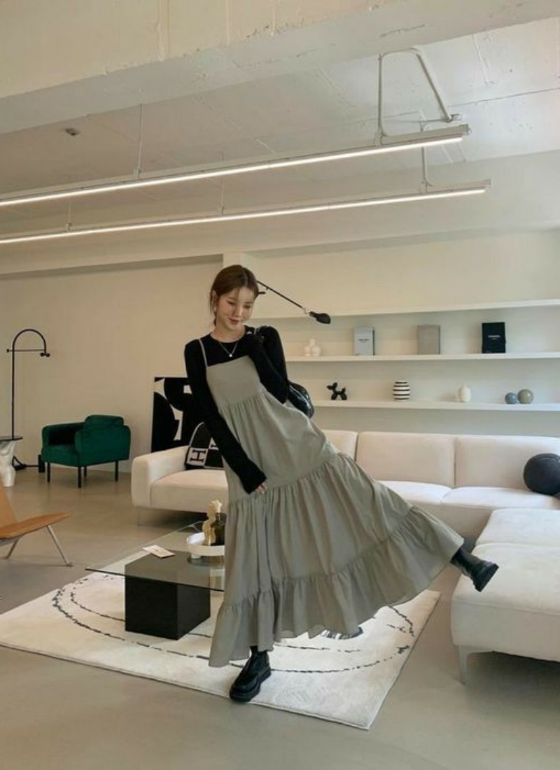 Người phụ nữ mặc váy dài màu xám đứng trong phòng khách hiện đại với ghế sofa và bàn trang trí.