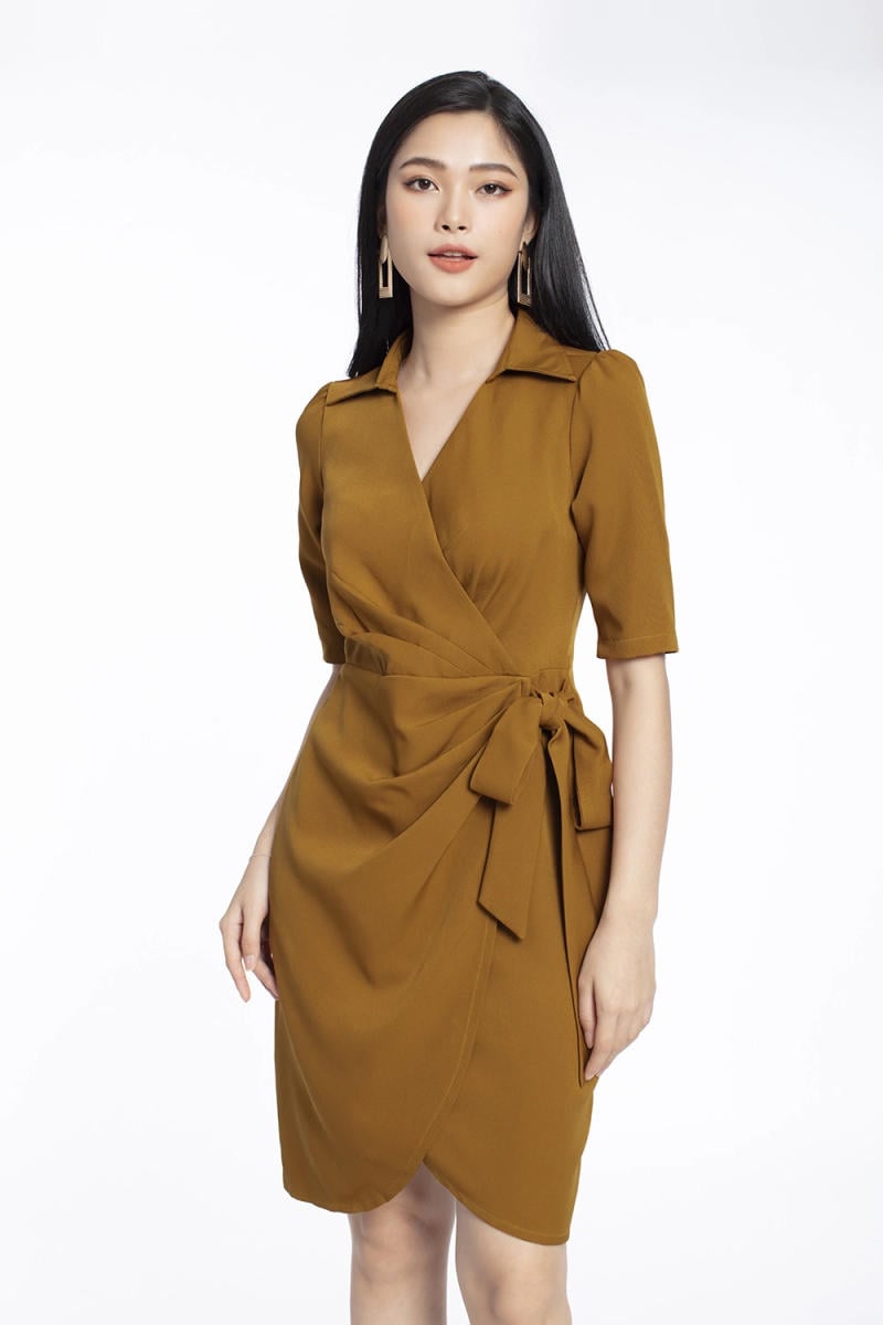 Người phụ nữ mặc váy màu nâu có thiết kế thắt nơ, tóc dài, đeo hoa tai, trên nền trắng.