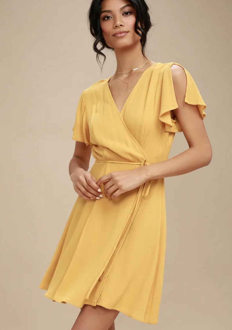 Người mặc váy vàng kiểu dáng quấn, tay ngắn xẻ, đứng trên nền màu be.