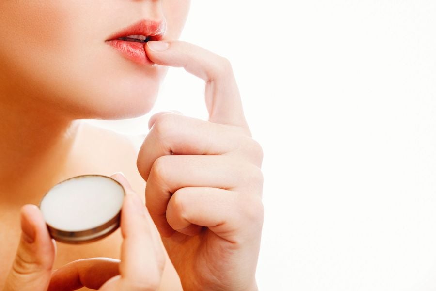 Người đang thoa son dưỡng lên môi bằng tay, cầm hộp son dưỡng nhỏ màu trắng.