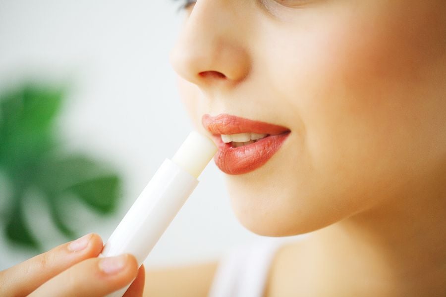 Người cầm thỏi son dưỡng môi gần môi, với nền lá cây mờ phía sau.