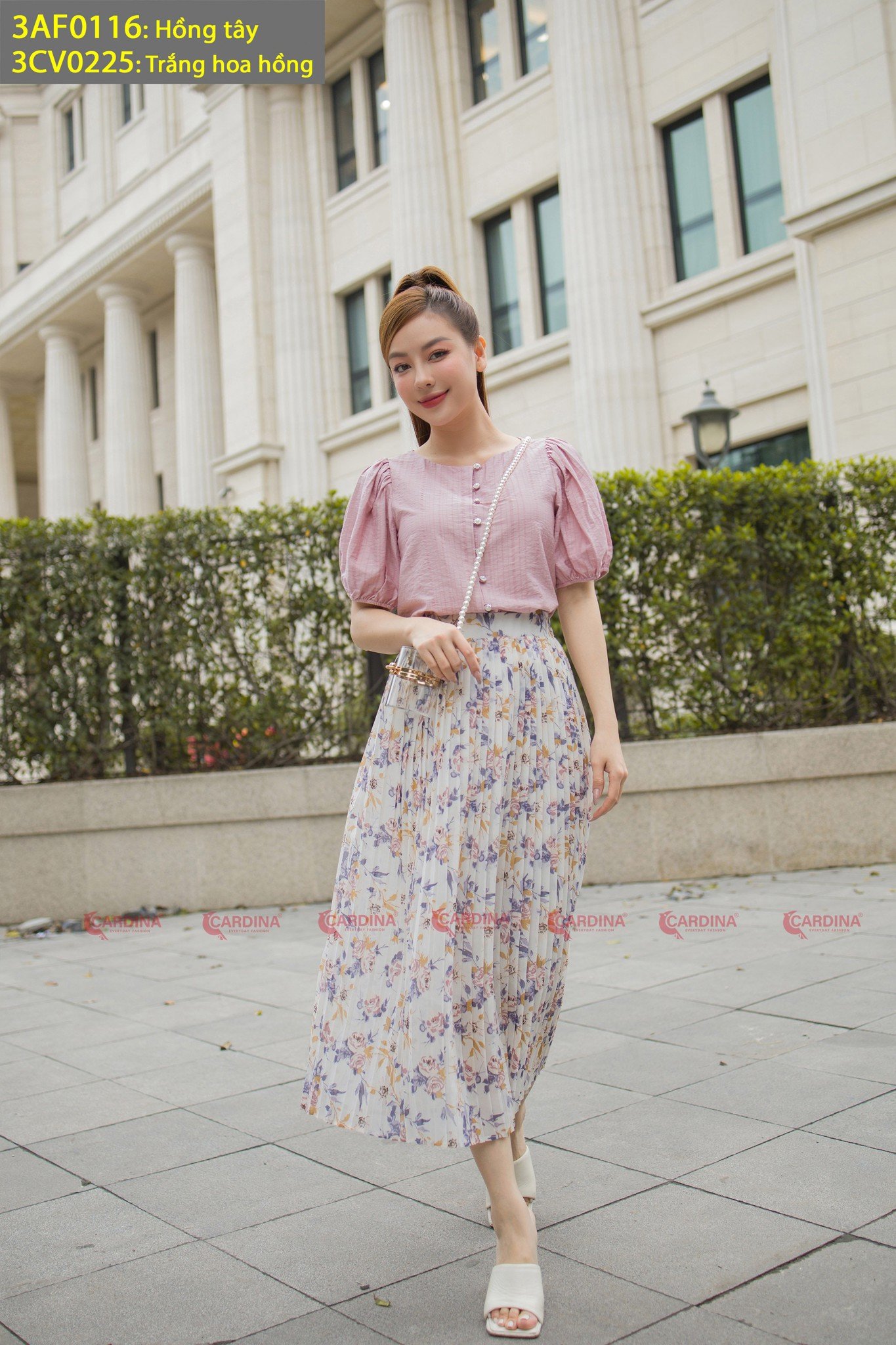 10 Cách Phối Đồ Với Chân Váy Hoa Nhí Giúp Bạn Tỏa Sáng Mùa Hè 18 Người phụ nữ mặc áo hồng và váy hoa dài đứng trước tòa nhà với cột và cửa sổ lớn.