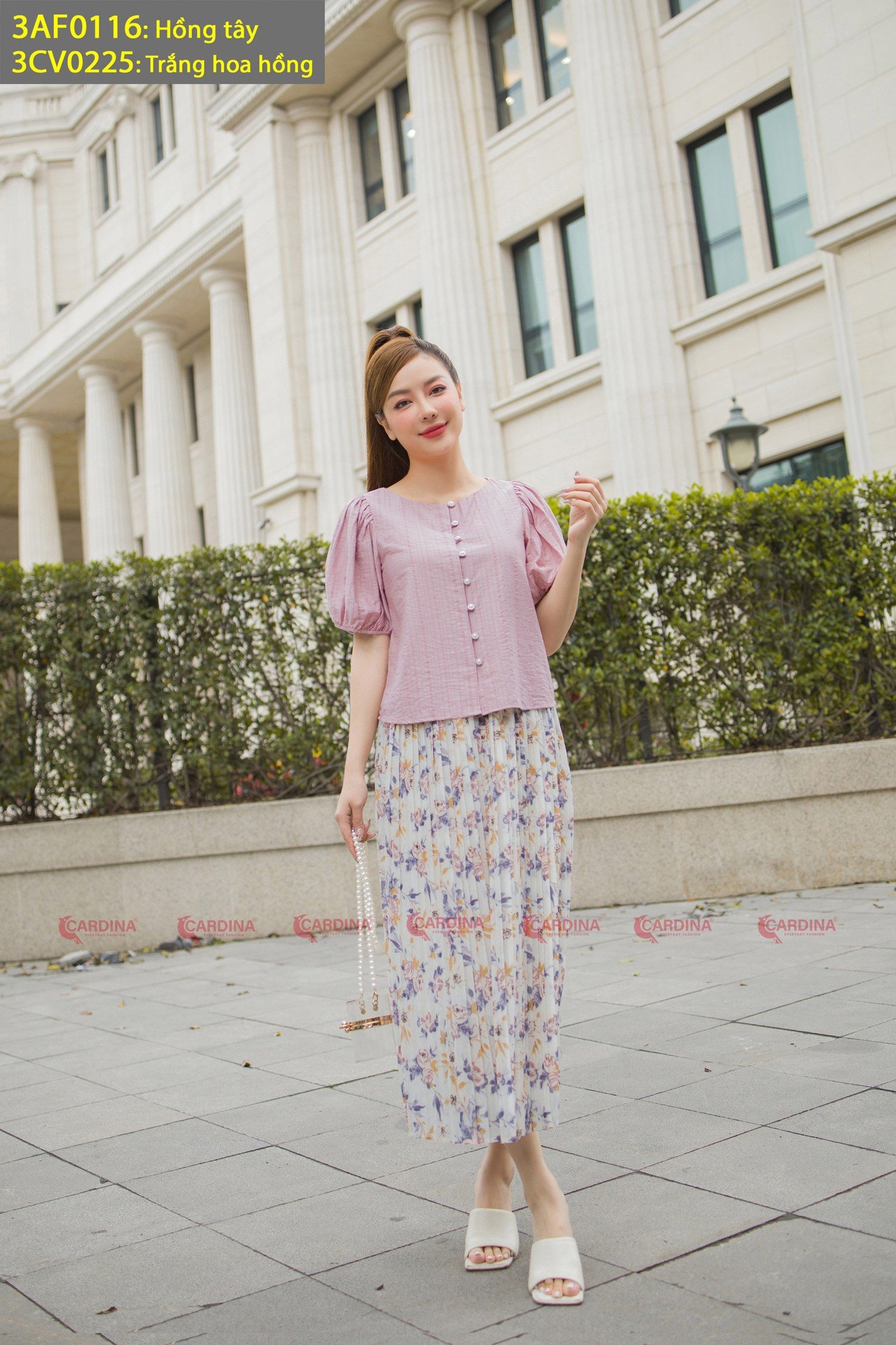 10 Cách Phối Đồ Với Chân Váy Hoa Nhí Giúp Bạn Tỏa Sáng Mùa Hè 19 Người phụ nữ mặc áo hồng và váy hoa đứng trước tòa nhà có cột lớn, tay cầm túi xách.