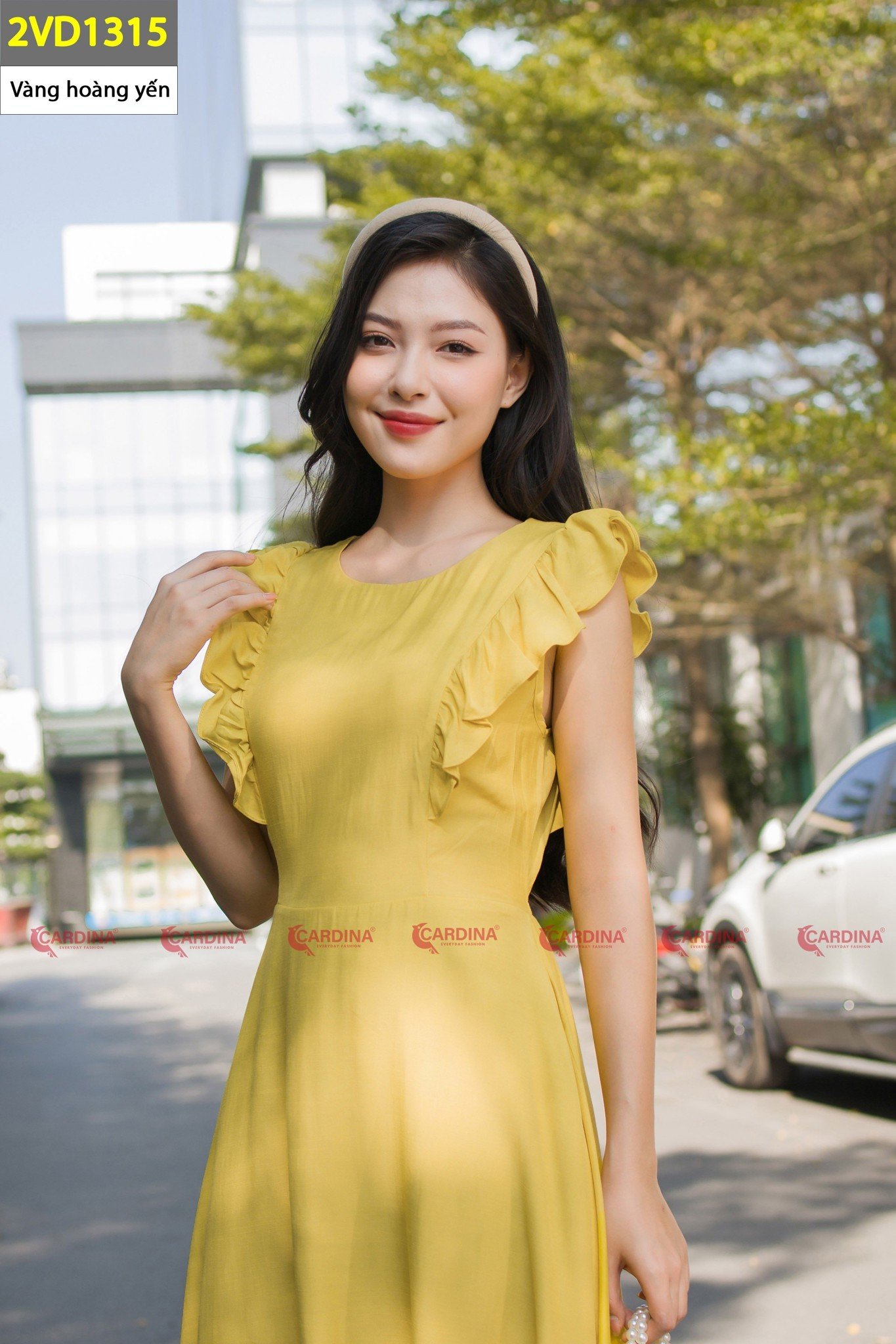 Người phụ nữ mặc váy vàng đứng ngoài trời, phía sau có cây xanh và tòa nhà hiện đại.