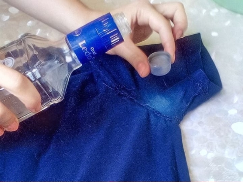 3fa21015-cach-tay-trang-quan-ao-10 Đang đổ chất lỏng từ chai lên một chiếc áo màu xanh, với nắp chai cầm trên tay.
