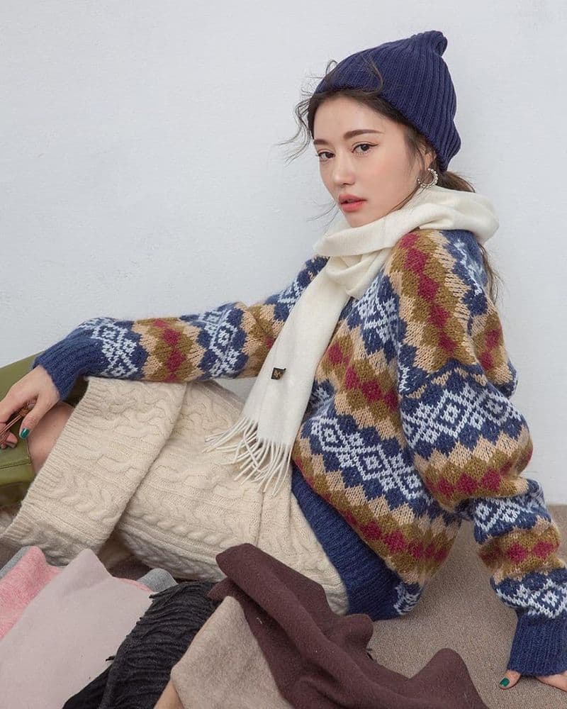 Người phụ nữ mặc áo len họa tiết, đội mũ len và quàng khăn trắng, ngồi trên nền sáng.