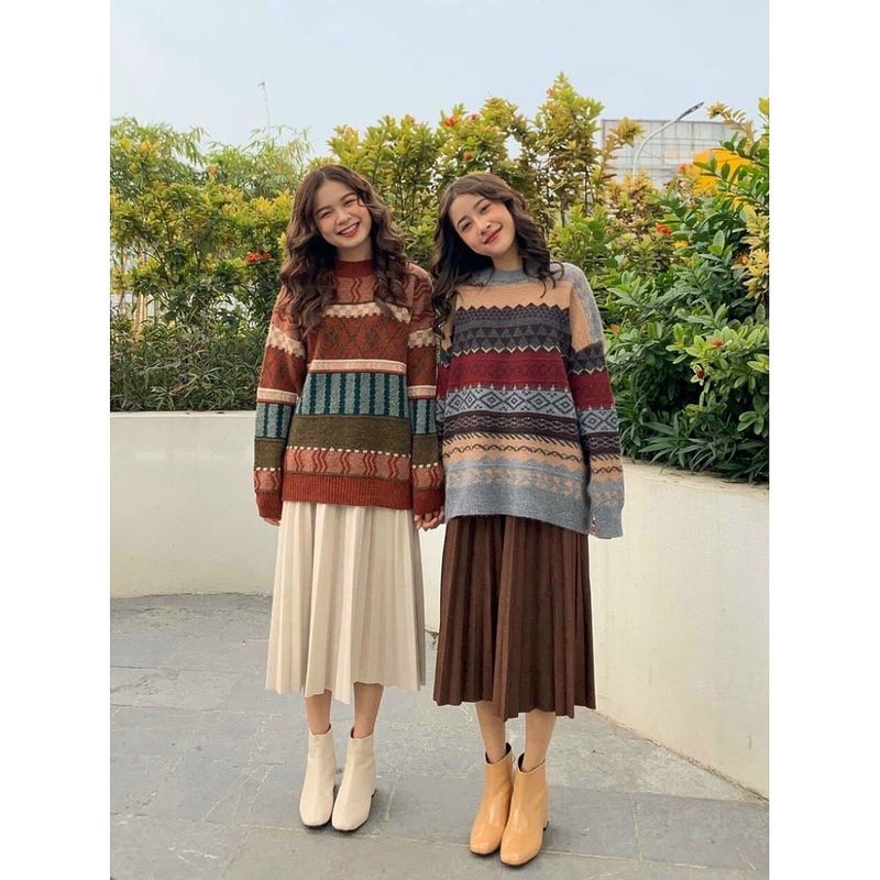 Hai người mặc áo len họa tiết và váy dài, đứng cạnh nhau trước bức tường trắng và cây cối xanh mướt.