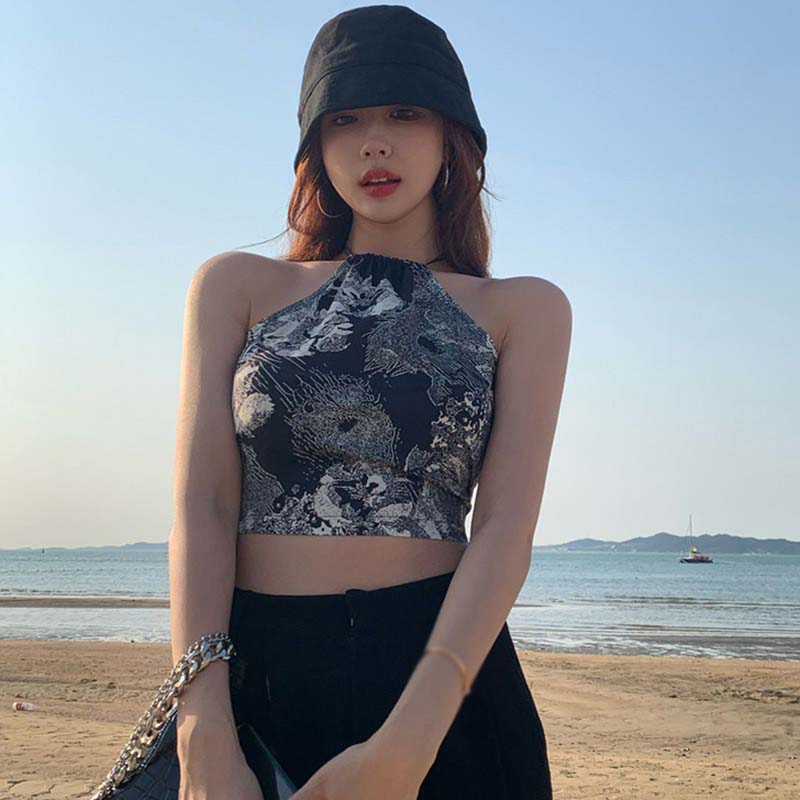 Người đội mũ đen đứng trước biển, mặc áo họa tiết, cầm túi xách, nền trời xanh và bãi biển.