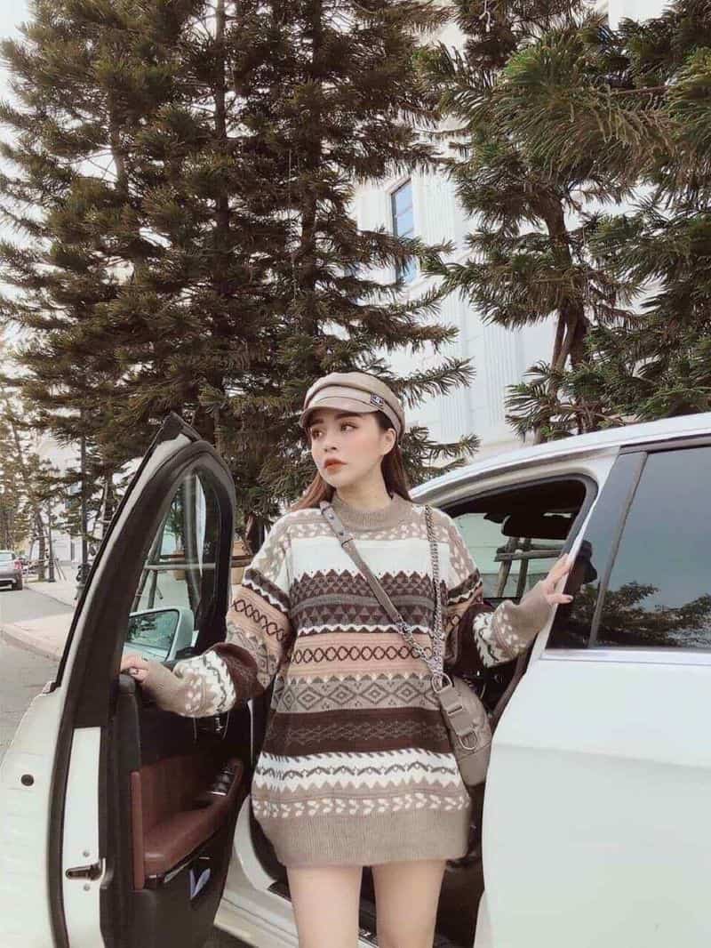 Người phụ nữ trong trang phục áo len họa tiết đứng bên cửa xe hơi mở, nền là cây xanh và tòa nhà.