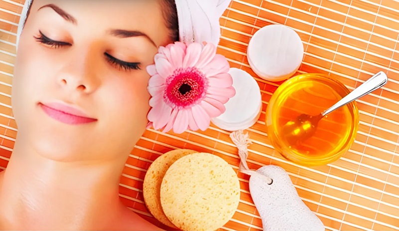 Người đang thư giãn với khăn tắm và hoa trên đầu, bên cạnh là mật ong và dụng cụ chăm sóc da.