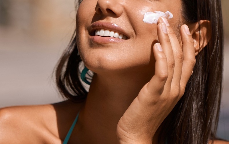 Người phụ nữ đang thoa kem dưỡng da lên mặt dưới ánh nắng, tươi cười và thoải mái.