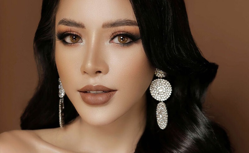 Người phụ nữ với trang điểm tinh tế, tóc đen dài, đeo hoa tai lấp lánh trên nền nâu.