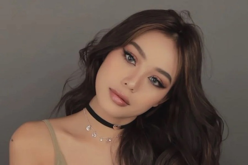 Người mẫu nữ có mái tóc dài, trang điểm kĩ lưỡng, đeo vòng cổ, nền màu xám.