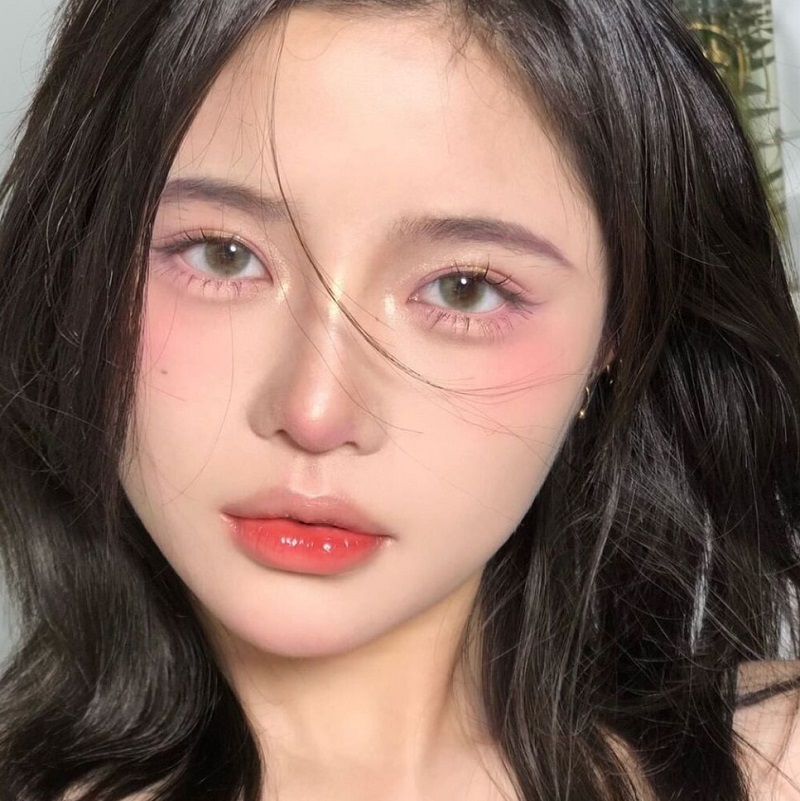 Người có mái tóc đen dài, trang điểm nhẹ nhàng với má hồng và môi đỏ, nhìn thẳng vào máy ảnh.