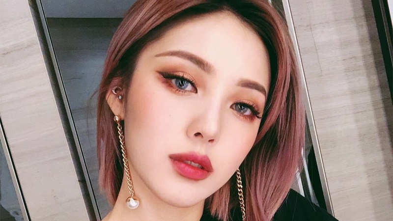 Người đang nhìn vào máy ảnh, tóc ngắn màu nâu, trang điểm mắt đậm, môi đỏ, đeo bông tai dài.