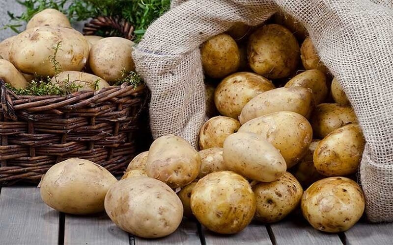 Nhiều củ khoai tây tươi rơi từ bao tải và nằm trên bàn gỗ, bên cạnh là giỏ mây cũng chứa đầy khoai tây.