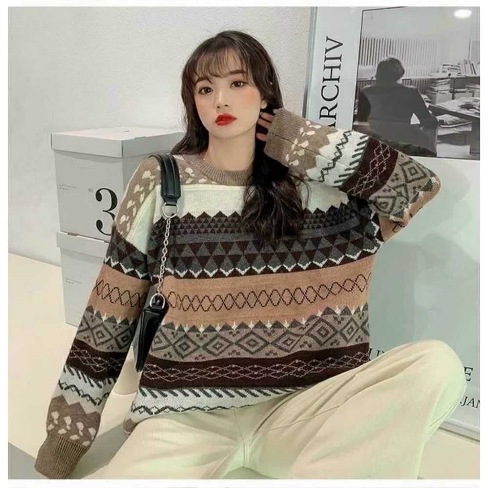 Người mặc áo len họa tiết đa dạng, tay cầm túi nhỏ, đứng trước tường trắng cạnh hộp số 3 và bức tranh đen trắng.
